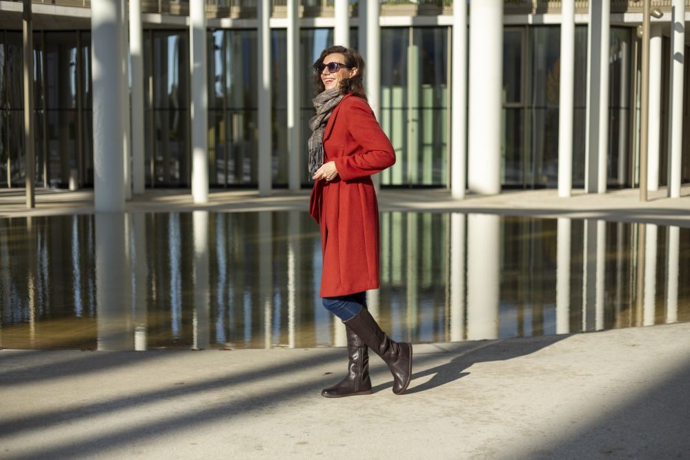 Eine Person in rotem Mantel, grauem Schal und Sonnenbrille schreitet selbstbewusst an einem runden Pool entlang, der von modernen weißen Säulen umgeben ist. Ihre QATCH Winter Brown-Stiefel von ZAQQ treten leichtfüßig auf, während das helle Sonnenlicht Schatten auf den Boden wirft.