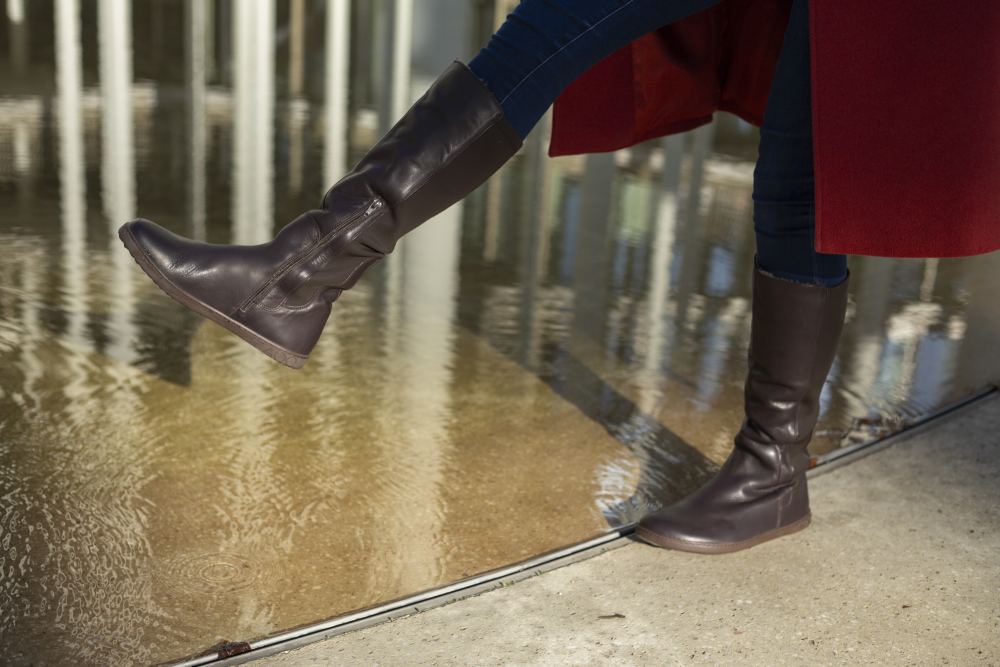 Eine Person in dunklen Jeans und einem Paar QATCH Winter Brown-Stiefeln von ZAQQ steigt in ein flaches Wasserbecken, teilweise bedeckt von einem langen roten Mantel. Die Spiegelung der umgebenden Strukturen im Wasser verleiht der Szene eine künstlerische Note.