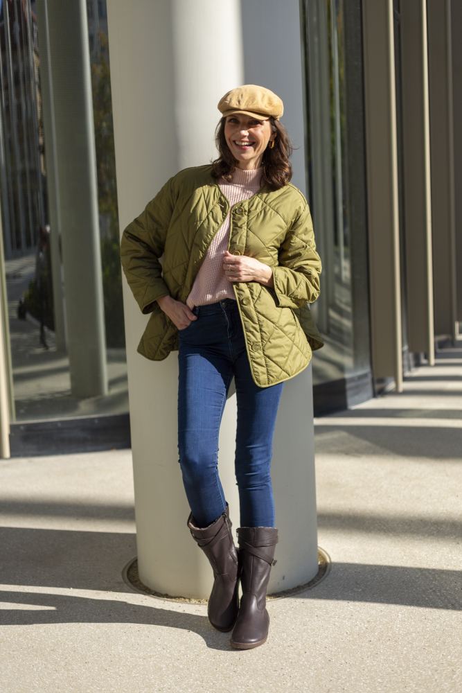 Eine Frau in grüner Steppjacke, rosa Pullover und Blue Jeans lehnt an einer Säule. Sie lächelt und trägt eine beige Mütze sowie ZAQQs ATTRAQT Winter Brown Stiefel für ultimativen Tragekomfort. Sonnenlicht wirft Schatten auf den Boden, im Hintergrund moderne Glaswände.