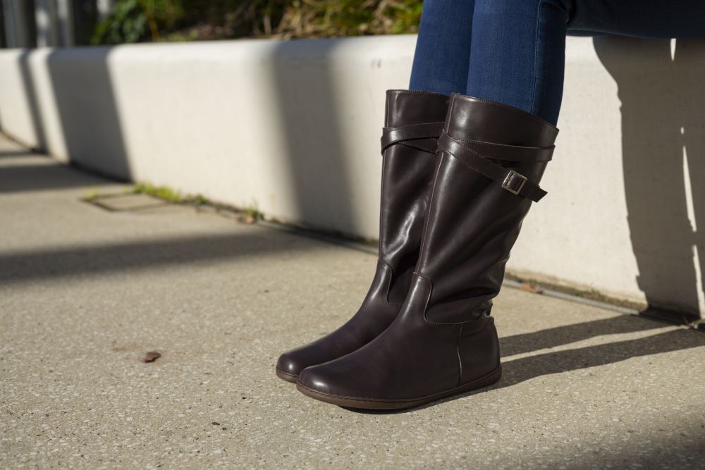 Nahaufnahme der Unterschenkel einer Person, die dunkelbraune ZAQQ ATTRAQT Nappa-Stiefel mit Schnallendetails trägt. Die Person sitzt auf einer niedrigen Betonmauer, Sonnenlicht wirft Schatten auf den Bürgersteig. In die Ziegenleder-Stiefel sind Jeans gesteckt.