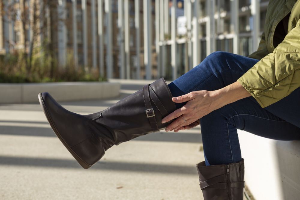 Im Freien sitzt eine Person in grüner Jacke und blauer Jeans und richtet ihre ATTRAQT Nappa Brown Boots von ZAQQ aus elegantem Ziegenleder. Im Hintergrund ist moderne Architektur mit Glas- und Metallelementen zu sehen.