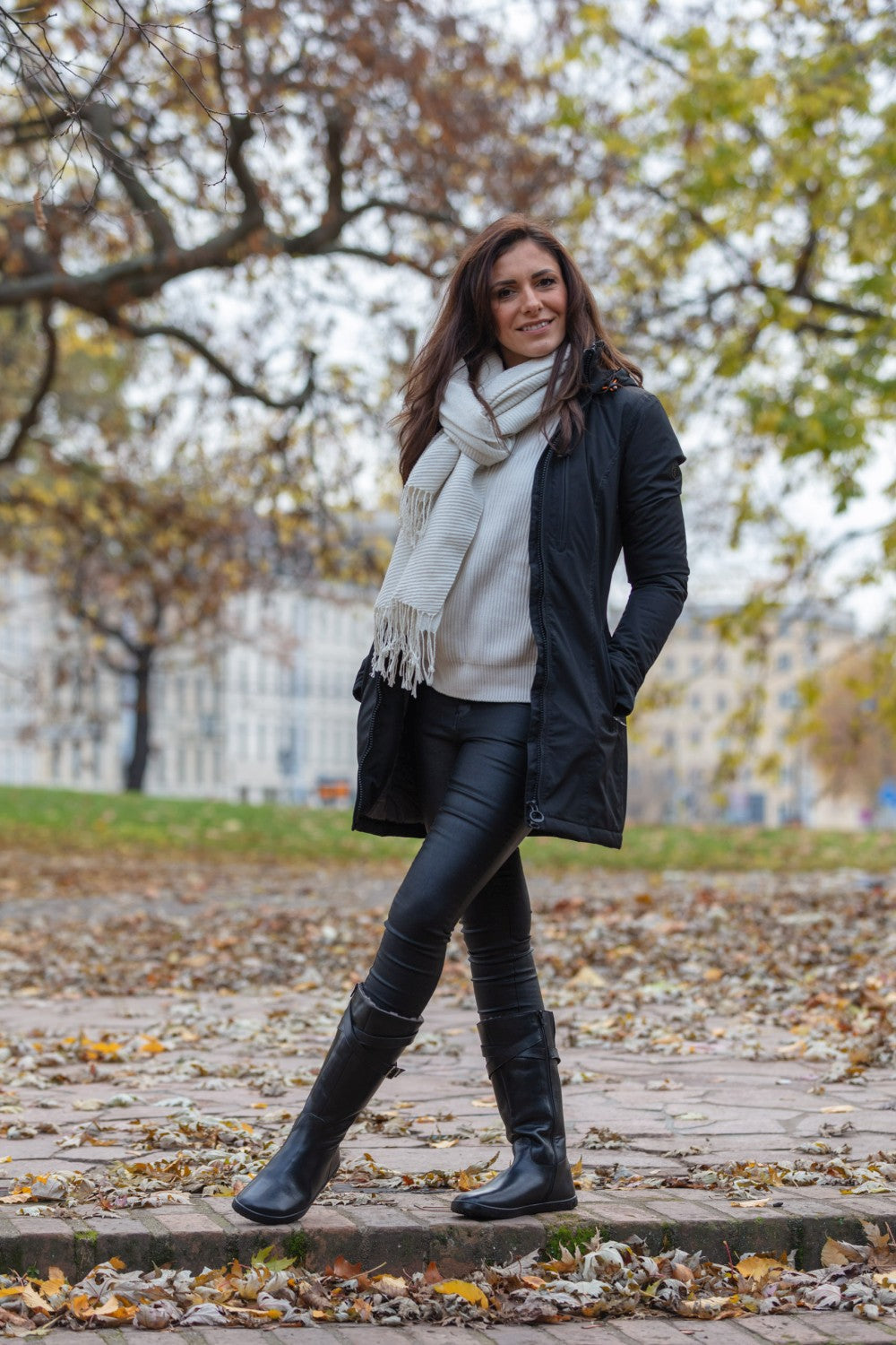 Eine Frau in weißem Pullover, Schal und schwarzem Mantel präsentiert ihren Stil mit ZAQQs ATTRAQT Winter Black Ziegennappaleder-Lederhose und Barfuß-Stiefel. Sie steht auf einem blätterbedeckten Weg, umgeben von Bäumen und Gebäuden, und fängt die Essenz der herbstlichen Landschaft ein.