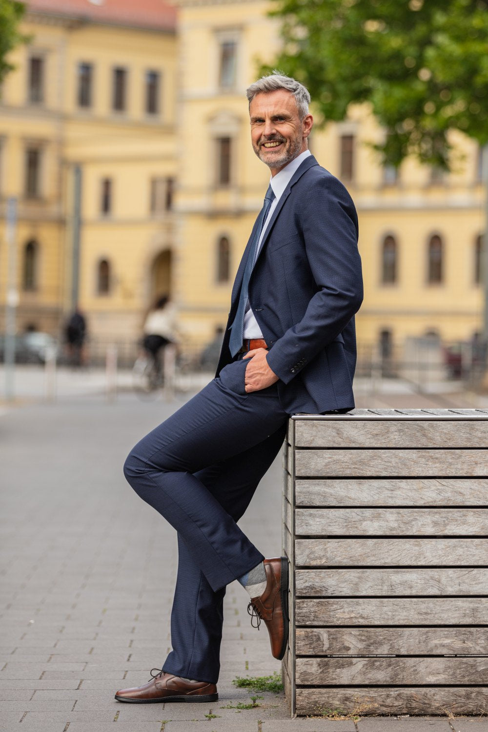 Ein Mann in einem marineblauen Anzug und braunen ZAQQ BRIQ Antique Brown-Schuhen lehnt an einer Holzkonstruktion in einer Stadtstraße. Er lächelt, im Hintergrund sind ein älteres Gebäude und Grünflächen zu sehen.