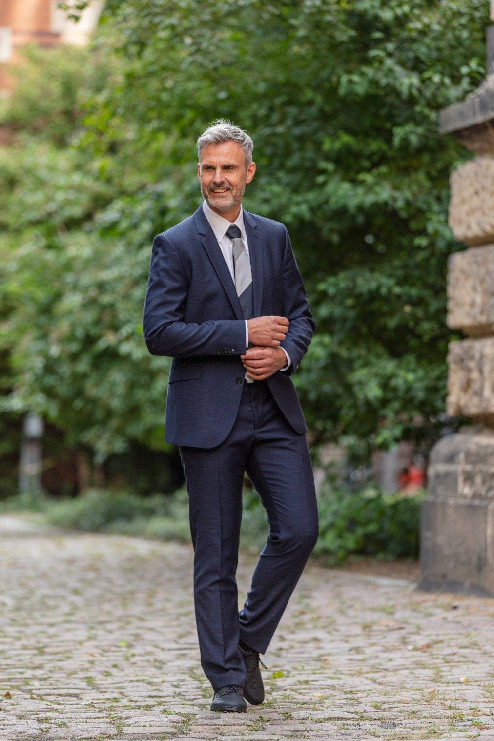 Ein Mann mit grauem Haar und Bart, gekleidet in einen dunkelblauen Anzug und Krawatte, geht gemütlich einen Kopfsteinpflasterweg inmitten üppigen Grüns entlang. Er trägt BRIQ Brogue Vegan Black Schuhe von ZAQQ mit rutschhemmender Sohle und zeigt ein kleines Lächeln und eine entspannte Haltung.