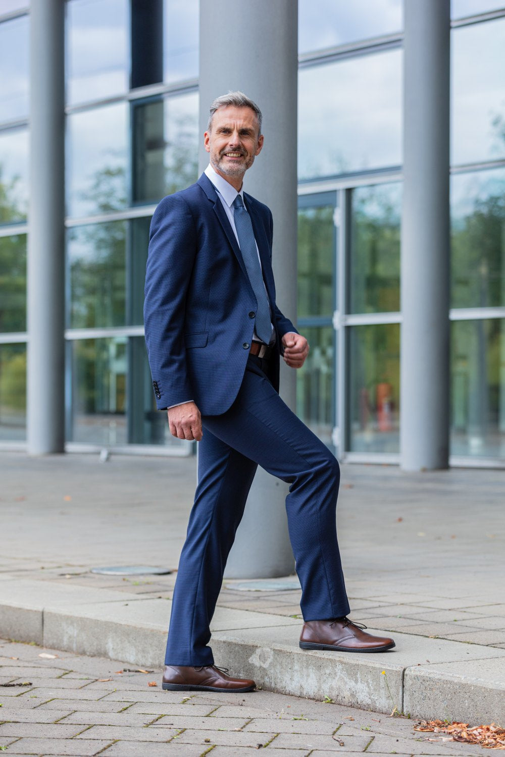 Ein Mann in blauem Anzug und weißem Hemd, der selbstbewusst die Schuhe DUQE Antique Brown von ZAQQ trägt, steht mit einem Fuß auf einer Stufe. Er steht im Freien vor einem modernen Glasgebäude. Der Himmel ist teilweise bewölkt und Herbstblätter liegen verstreut auf dem Boden.