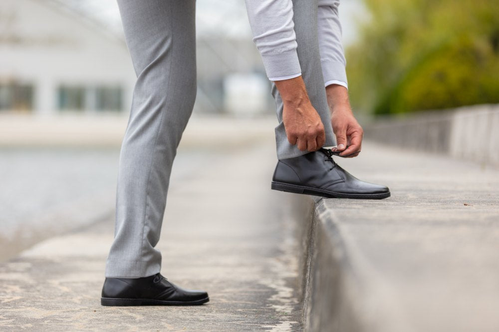 Eine Person in hellgrauen Hosen und schwarzen DUQE-Schuhen von ZAQQ hockt auf einer Betonfläche, um ihre Schnürsenkel zu binden, mit einem verschwommenen Hintergrund aus Grün und einem Gebäude in der Ferne.