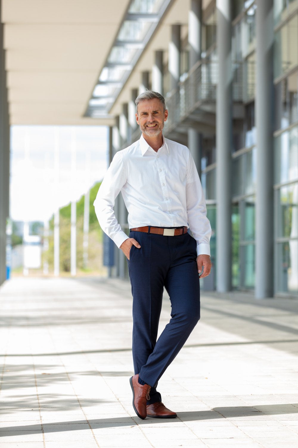 Ein selbstbewusster Mann in weißem Hemd und dunkler Hose steht mit den Händen in den Taschen in einem modernen Außenkorridor. Sein leichtes Lächeln passt gut zu den eleganten PEAQ Cognac-Schuhen von ZAQQ, die er trägt, während er lässig an einer Säule lehnt. Im Hintergrund sind Glasfenster und ein klarer Himmel zu sehen.