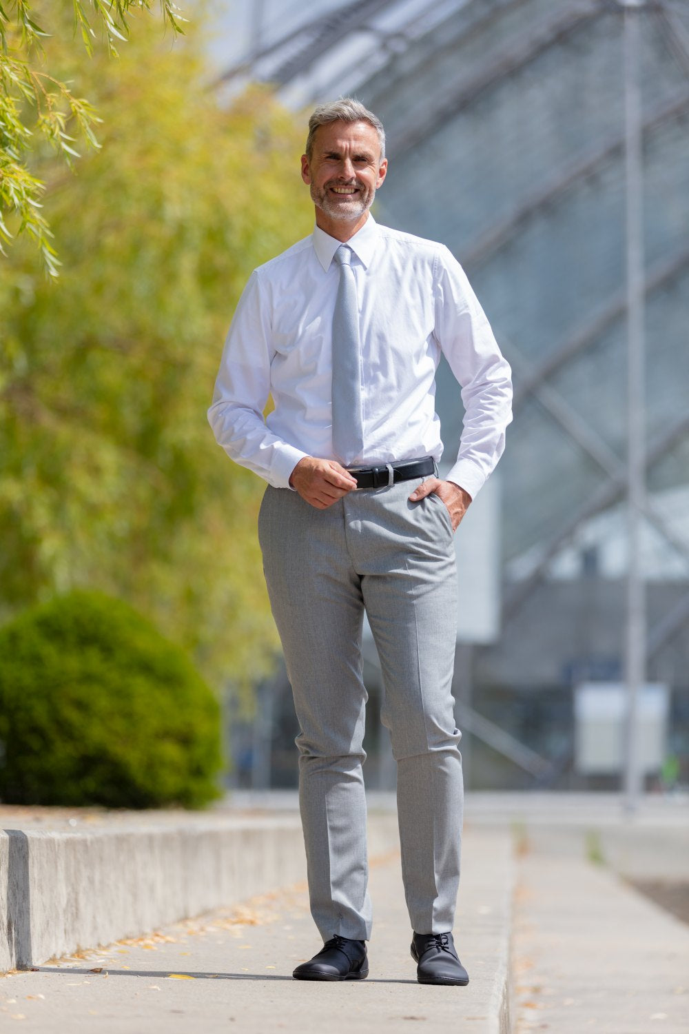 Ein Mann mit grauem Haar und Bart steht draußen in einer modernen, urbanen Umgebung. Er trägt ein weißes Hemd, hellgraue Anzughosen und elegante PEAQ Vegan Black-Schuhe von ZAQQ. Er lächelt vor einer Kulisse aus Glasarchitektur und Grün. Seine Schuhe sind mit atmungsaktivem Mikrofaserfutter für mehr Komfort ausgestattet.