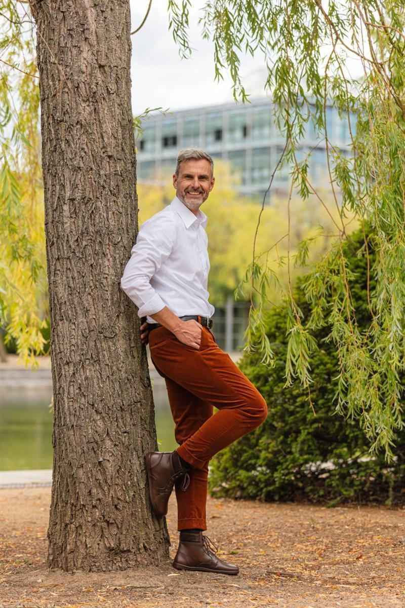 Ein Mann mit grauem Haar und Bart lehnt an einem Baum. Er trägt ein weißes Hemd, braune Hosen und robuste QORBY Brown-Stiefel von ZAQQ. Der Hintergrund aus Bäumen, einem See und einem Gebäude fängt die Essenz eines entspannten Herbsttages im Freien ein.