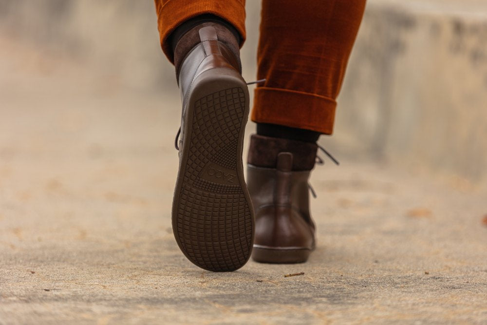 Eine Nahaufnahme zeigt eine Person, die auf einem Bürgersteig geht. Sie zeigt ein Paar braune ZAQQ QORBY Halbhohe Stiefel aus Leder, kombiniert mit einer orangefarbenen Hose. Der Fokus liegt auf der Sohle eines Stiefels, die ihr strukturiertes Griffmuster hervorhebt, während der Hintergrund unscharf bleibt.