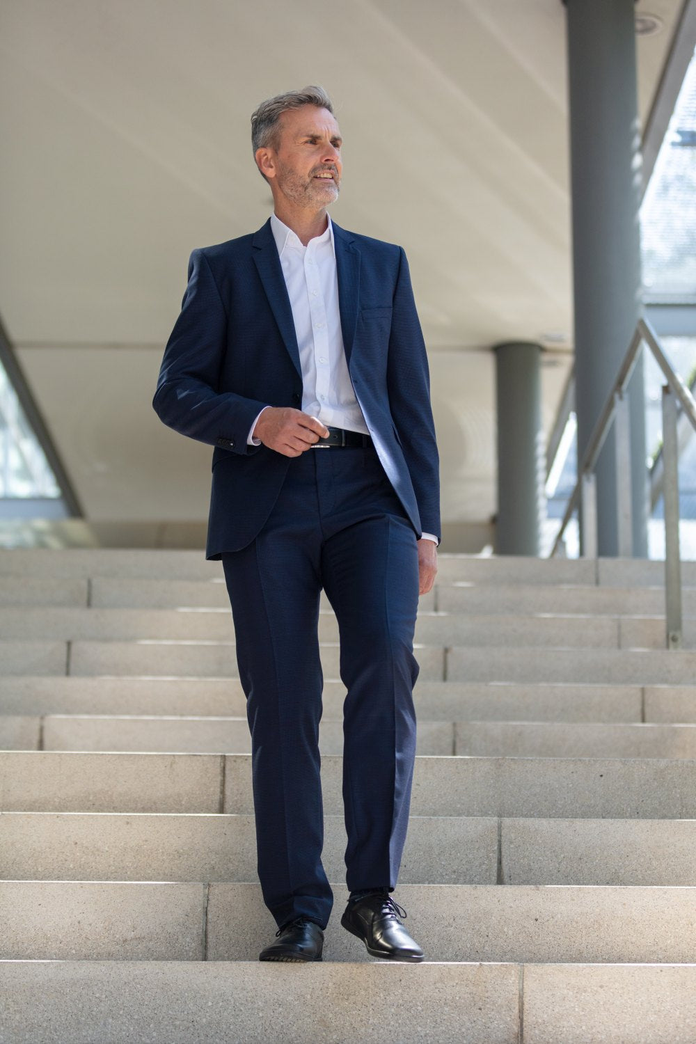 Ein Mann in blauem Anzug und weißem Hemd schreitet selbstbewusst eine graue Treppe hinunter, seine schwarzen SEQUENT-Schuhe von ZAQQ glänzen bei jedem Schritt. Er blickt mit einem leichten Lächeln geradeaus, das Telefon in der Hand. Die moderne Architektur um ihn herum zeichnet sich durch große Säulen und Glas aus, was sein selbstbewusstes Auftreten ergänzt.