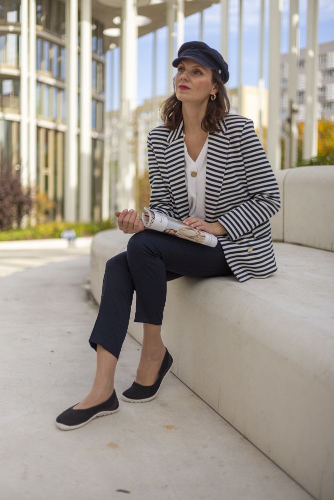 Eine Frau sitzt auf einer modernen Steinbank im Freien und hält eine Zeitschrift in der Hand. Sie trägt einen gestreiften Blazer, ein weißes Oberteil, eine dunkle Hose und SUQAR schwarze Barfußschuhe von ZAQQ für die perfekte Mischung aus Komfort und femininem Look. Eine marineblaue Kappe rundet ihren Stil vor der Kulisse zeitgenössischer Architektur mit Glas- und Metallelementen ab.