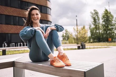 Eine Frau sitzt draußen auf einer Holzbank und lächelt in die Kamera. Sie trägt ein blaugrünes Outfit und leuchtend orangefarbene Turnschuhe. Ihr Haar weht im Wind. Im Hintergrund sind ein modernes Gebäude und etwas Grün unter einem bewölkten Himmel zu sehen.