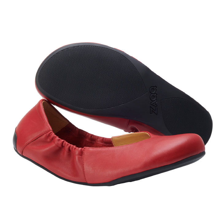 Ein Paar MAIQA Red Ballerinas von ZAQQ hat eine schwarze Sohle. Ein Schuh steht aufrecht und betont die elastische Rückseite und das weiche Nappaleder, während der andere darüber steht und die gemusterte Sohle mit dem geprägten ZAQQ-Logo enthüllt.