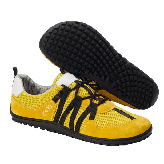 Ausgestellt sind RIQ Yellow-Sportschuhe von ZAQQ mit schwarzen Sohlen und Schnürsenkeln für ein flexibles Tragegefühl. Der Markenname ist deutlich sichtbar auf die Seite gedruckt und ein Schuh ist umgedreht, um das für Barfußschuhe typische strukturierte Griffmuster zu zeigen.