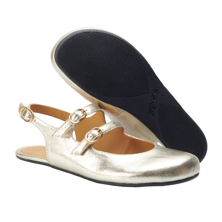 Ein Paar Ballerinas im Mary-Jane-Stil von ZAQQ QOMET Gold versprühen mit ihrem glänzenden Finish Glamour, verfügen über zwei Schnallenriemen und flexible schwarze Sohlen. Ein Schuh steht aufrecht, während der andere umgedreht ist, um die Haltbarkeit seiner Sohle zu präsentieren.