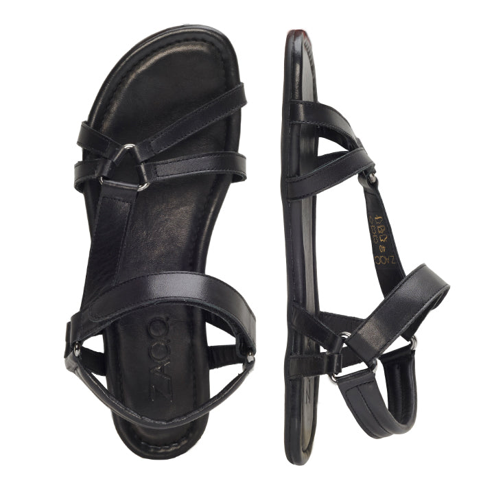 Ein Paar schwarze QALA-Sandalen von ZAQQ aus hochwertigem Nappaleder mit dünnen Riemen und einer flachen, rutschfesten Sohle. Die linke Sandale ist mit der Sohle nach oben abgebildet, während die rechte eine elegante Seitenansicht bietet.