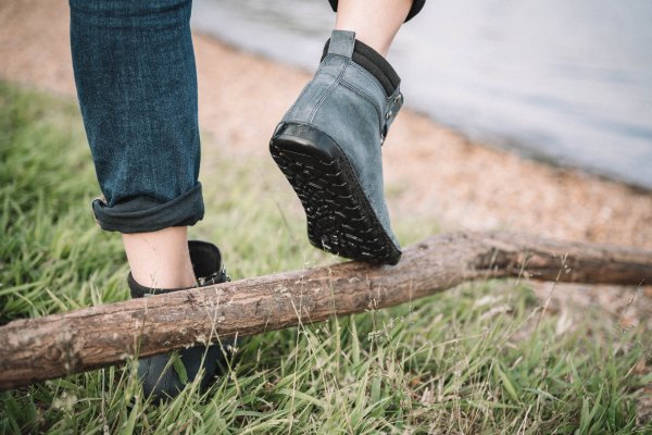 Eine Person in hochgekrempelten Jeans und schwarzen Stiefeletten balanciert auf einem umgestürzten Ast. Die Stiefel haben ein sichtbares Profilmuster. Die Kulisse besteht aus Grasland und einem Kieselstrand neben einem Gewässer, was auf eine Umgebung im Freien hindeutet.