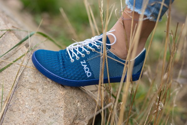 Der Fuß einer Person steht auf einem großen Felsen und trägt einen blauen gewebten Sneaker mit weißen Schnürsenkeln und einer marineblauen Sohle, der mit der Marke ZAQQ versehen ist. Der Sneaker besteht aus einem strukturierten, atmungsaktiven Material. Die Person trägt hochgekrempelte hellblaue Jeans. Trockenes Gras umgibt den Felsen in einer natürlichen Umgebung im Freien.