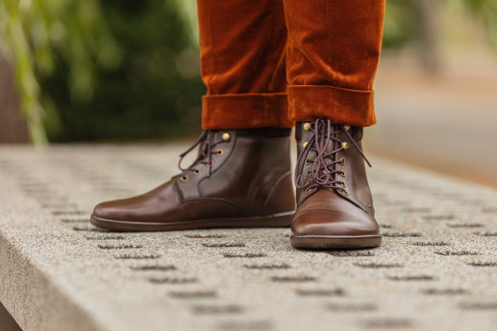 Eine Person, die braune Lederstiefel mit Schnürung und orangefarbene Cordhosen trägt, steht auf einer strukturierten Betonfläche. Die Stiefel haben goldene Ösen und dunkelbraune Schnürsenkel. Die Hosen sind hochgekrempelt, sodass die Stiefel vollständig sichtbar sind. Der Hintergrund ist unscharf und lässt auf eine Außenkulisse schließen.