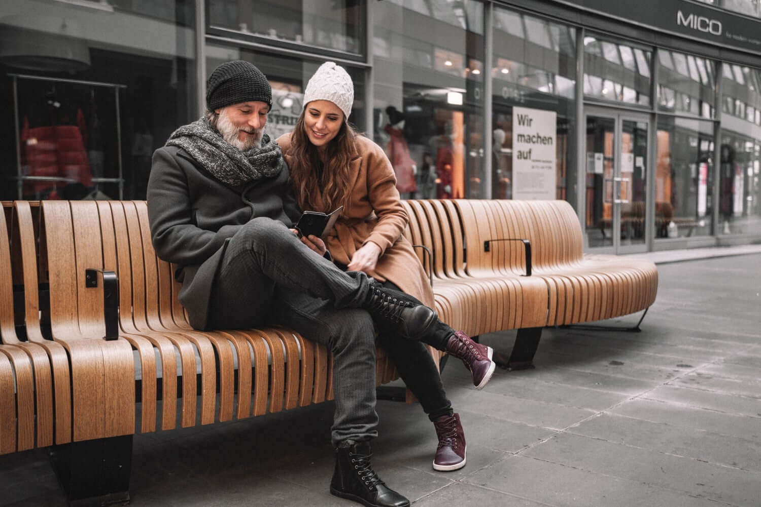 Ein Mann und eine Frau sitzen auf einer modernen Holzbank. Beide tragen Wintermäntel und Strickmützen. Der Mann mit dem grauen Bart schaut auf ein Smartphone, während die Frau lächelt und sich nah zu ihm beugt. Sie befinden sich in einem überdachten Einkaufsbereich mit Ladenschildern und Menschen im Hintergrund.