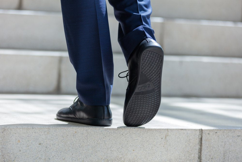 Eine Person in einer dunkelblauen Hose und schwarzen ARQ-Schuhen von ZAQQ läuft Betonstufen hinauf. Der Fokus liegt dabei auf der Sohle, die das elegante Profil des Glattleders zur Geltung bringt.