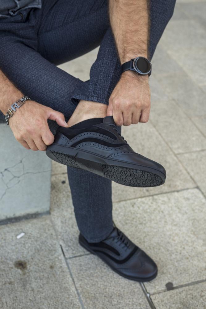 Eine Person in grauer Hose und ZAQQs BRIQ Brogue Deep Black-Schuhen sitzt auf einer Betonfläche und richtet einen Schuh. Die elegante schwarze Armbanduhr ergänzt ein Armband an ihrem Handgelenk, wobei das Bild ihre Hände und die lässige Eleganz der Barfußschuh-Sneaker hervorhebt.