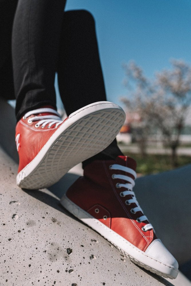 Eine Person trägt ZAQQs rote CHUQQS High-Top-Sneaker mit weißen Schnürsenkeln und sitzt im Freien auf einer glatten Betonfläche. Der Fokus liegt auf den Schuhen, die wie Barfußschuhe bequem gestaltet sind, mit einem verschwommenen Baum und blauem Himmel im Hintergrund.