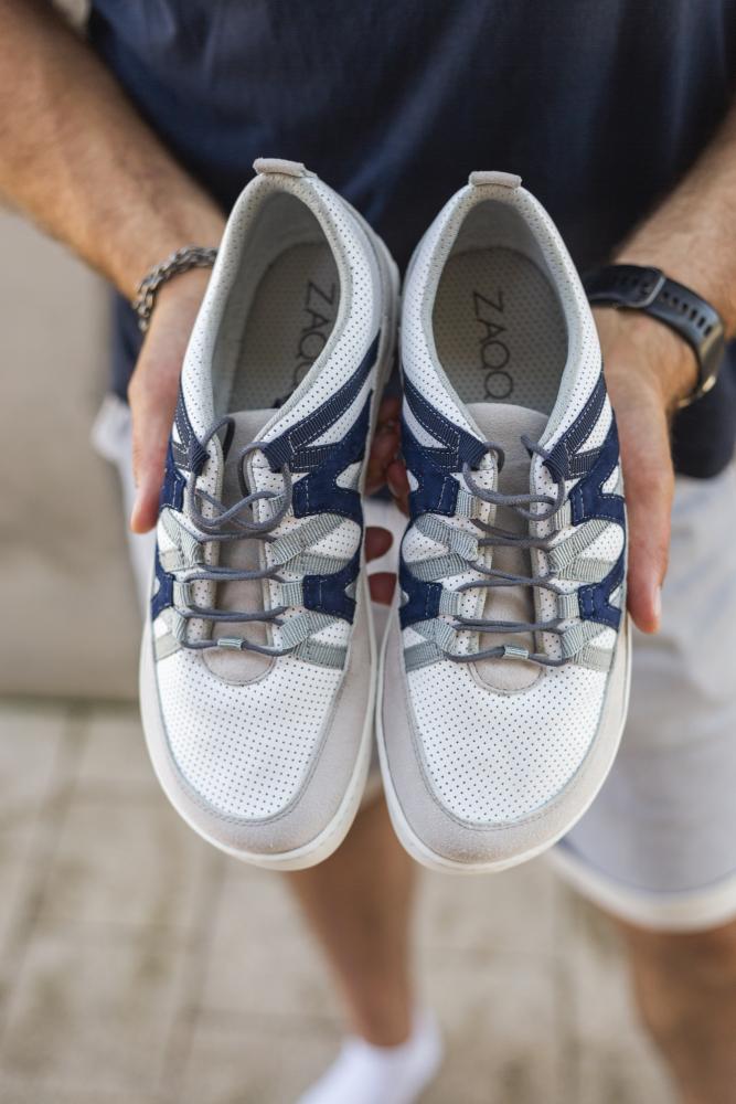 Eine Person hält ein Paar DRAQE White Grey-Sneaker von ZAQQ in den Farben Weiß und Marineblau mit grauen Akzenten. Diese Schuhe sind aus hochwertigem Nappaleder gefertigt und weisen ein strukturiertes Design sowie Ösen für Schnürsenkel auf. Der leicht verschwommene Hintergrund unterstreicht ihren Barfußschuh-Stil.