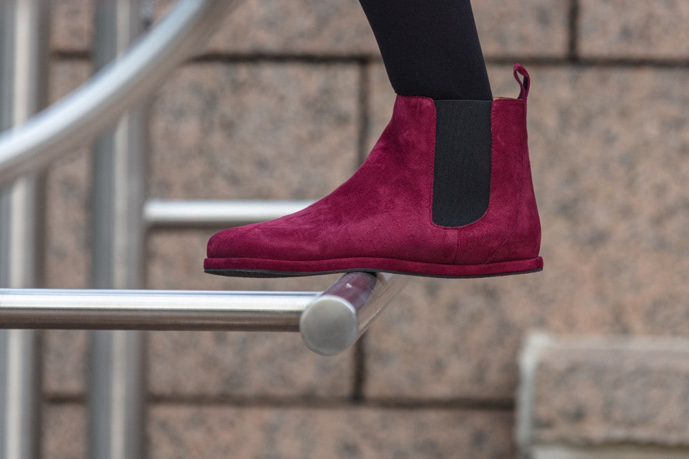 Eine Person in schwarzen Leggings und auffälligen ZAQQ EQUITY Berry-Stiefeln aus leuchtend rotem Wildleder steht auf einem silbernen Geländer, im Hintergrund eine Steinmauer, die einen Kontrast zu ihrer gewagten Schuhwahl bildet.