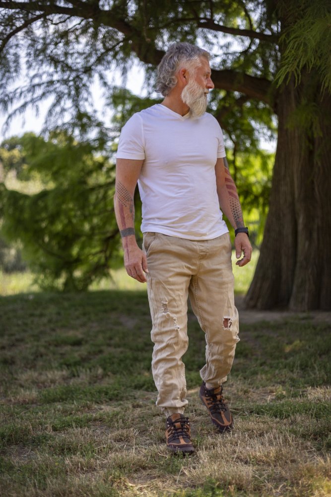 Ein Mann mit weißem Bart und Tattoos läuft auf Gras. Er trägt ein weißes T-Shirt, beige zerrissene Hosen und braune ESQAPE Waterproof-Schuhe von ZAQQ. Die Szene im Freien wird durch grüne Bäume im Hintergrund vervollständigt.