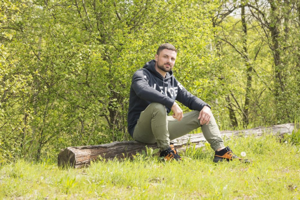 Ein Mann in einem dunklen Kapuzenpullover und grünen Hosen sitzt auf einem großen Baumstamm in einer von üppigen grünen Bäumen umgebenen Grasfläche und trägt die wasserdichten Schuhe ESQAPE Black Orange von ZAQQ. Er wirkt entspannt, ein Arm ruht unter dem klaren Himmel auf seinem Knie.