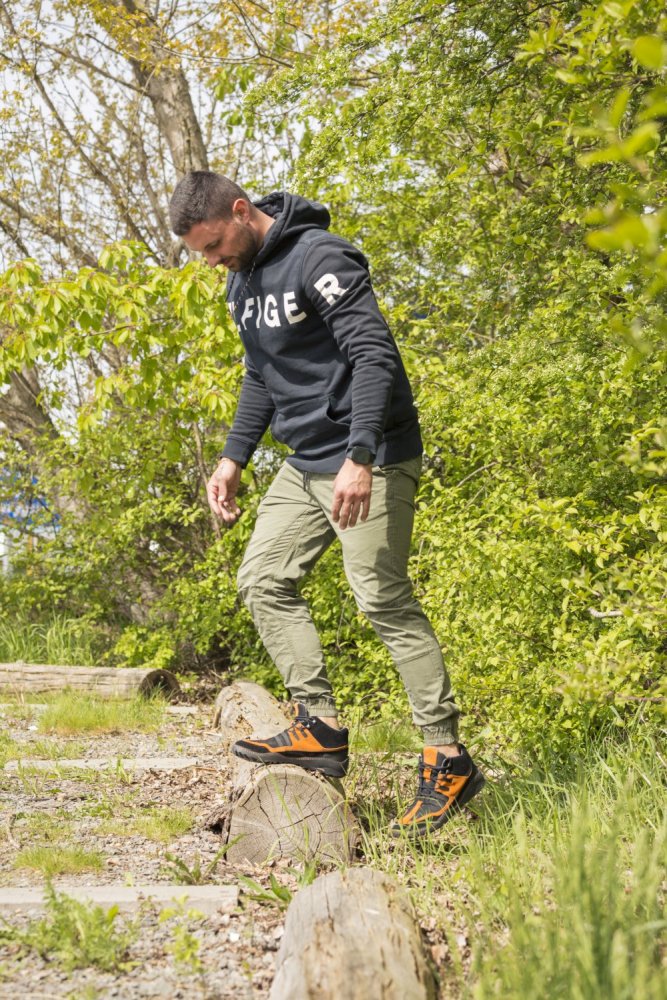 Ein Mann in einem schwarzen Kapuzenpullover und grünen Hosen geht vorsichtig auf Holzstufen, die von grünem Laub umgeben sind. Seine Füße sind bequem in die wasserdichten Schuhe ESQAPE Black Orange von ZAQQ gekleidet, was für ein natürliches Gefühl sorgt.