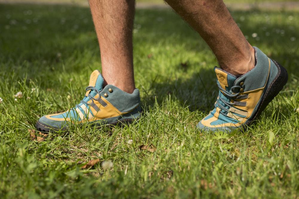 Eine Person läuft auf Gras und trägt wasserdichte ESQAPE-Wanderschuhe in Blau und Orange von ZAQQ. Der Fokus liegt auf dem wasserdichten Schuhwerk und den Unterschenkeln, mit einem verschwommenen Hintergrund aus grünem Gras und kleinen weißen Blumen.