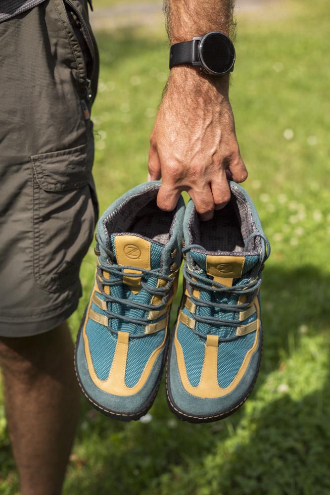 Eine Person in Shorts hält ein Paar blau-orangefarbene Wanderschuhe, die ESQAPE Blue Orange Waterproof von ZAQQ, über eine Grasfläche. Die Armbanduhr der Person ist sichtbar.