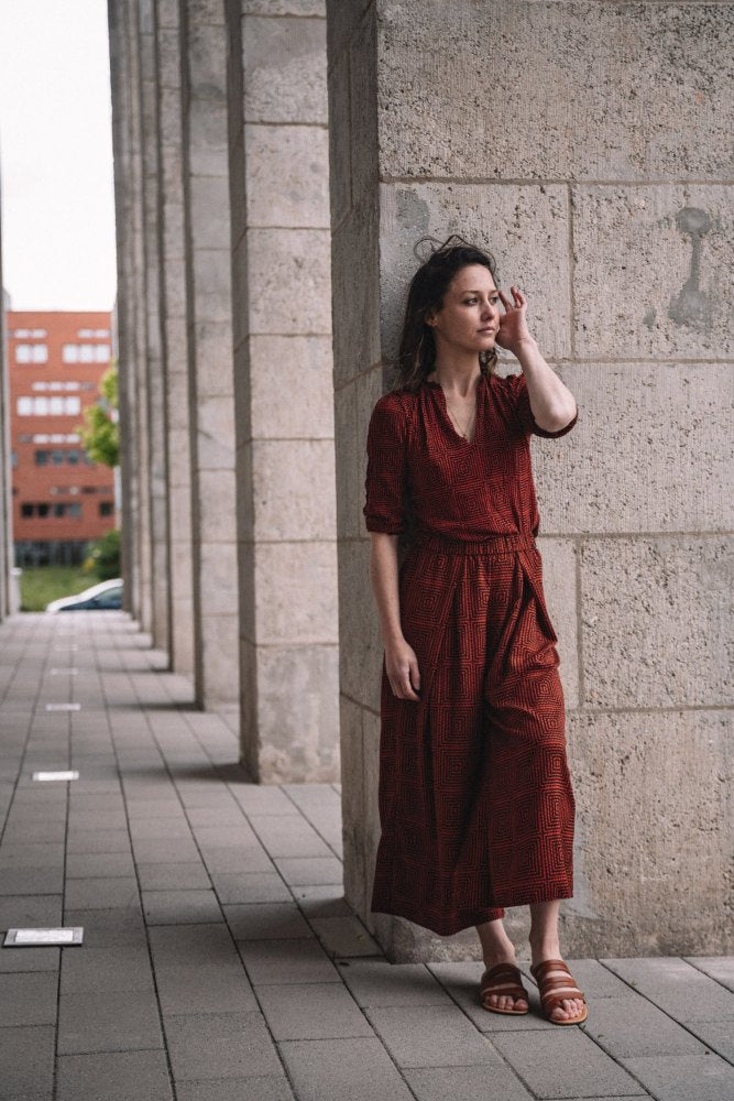 Eine Frau in einem langen, roten Kleid und FLAQE Brown Sandalen von ZAQQ steht an einer Steinmauer auf einem von Säulen gesäumten Gehweg. Sie berührt ihr Haar und schaut zur Seite. Im Hintergrund ist ein Gebäude mit roten Ziegeln und Grün zu sehen.