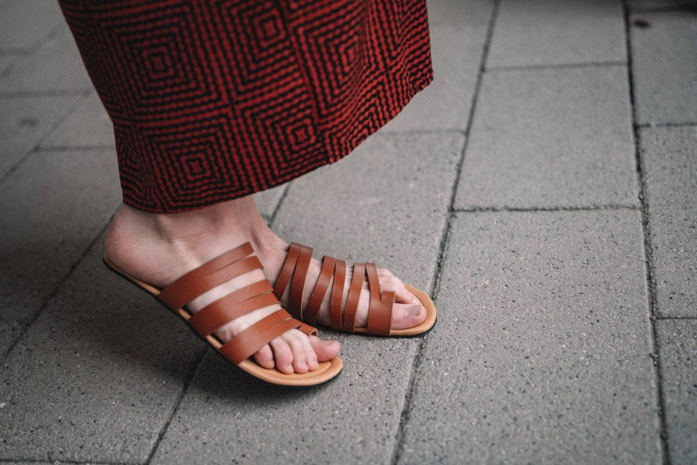 Eine Person, die braune FLAQE-Sandalen von ZAQQ und einen rot gemusterten Rock trägt, steht auf einem grau gefliesten Bürgersteig.