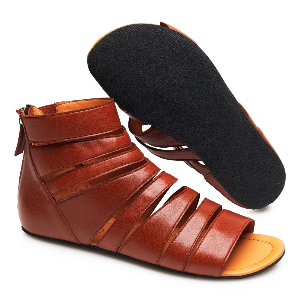 Ein Paar GLADIQ Cognac-Sandalen von ZAQQ, wobei eine Sandale von der Seite und die andere von unten gezeigt wird. Diese braunen Sandalen im Gladiator-Stil haben mehrere Riemen, einen Reißverschluss auf der Rückseite und eine schwarze flache Sohle.