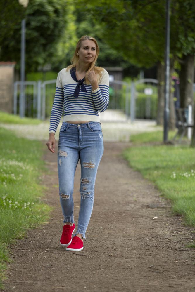 Jemand läuft einen Feldweg im Park entlang. Er trägt einen gestreiften Pullover, zerrissene Jeans und auffällige LIQE Race-Sneaker von ZAQQ. Die Szene spielt sich vor einem Hintergrund aus üppigem grünem Gras und Bäumen ab, mit einem Zaun und einem Weg, der sich in die Ferne erstreckt.