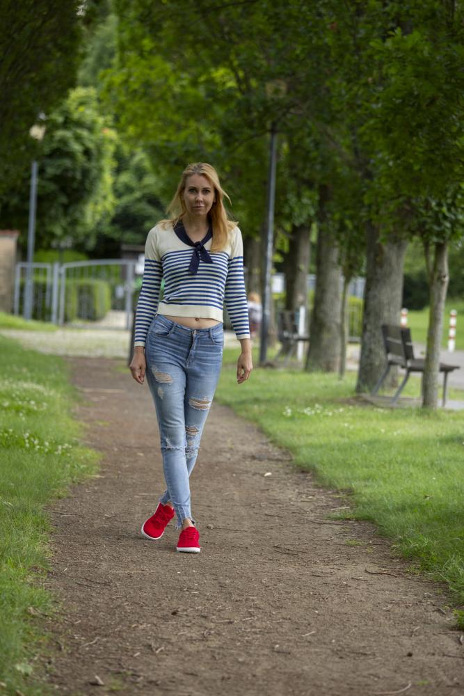 Eine Frau mit langen blonden Haaren schlendert einen von Bäumen gesäumten Weg entlang. Sie trägt ein gestreiftes Oberteil und zerrissene Jeans. Selbstbewusst tritt sie in einem Paar leuchtend roter ZAQQ LIQE Race-Sneaker aus Merinowolle auf den Weg. Entlang des Weges sind Bänke und Grasflächen zu sehen.