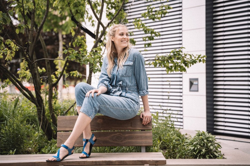 Eine Frau im Jeans-Outfit sitzt auf einer Holzbank im Freien, lächelt und blickt zur Seite. Sie ist von Bäumen und Grün umgeben, im Hintergrund ist ein modernes Gebäude zu sehen. An ihren Füßen trägt sie die MIQA Blue-Sandalen von ZAQQ mit Klettverschlüssen, die sowohl Komfort als auch Stil bieten.