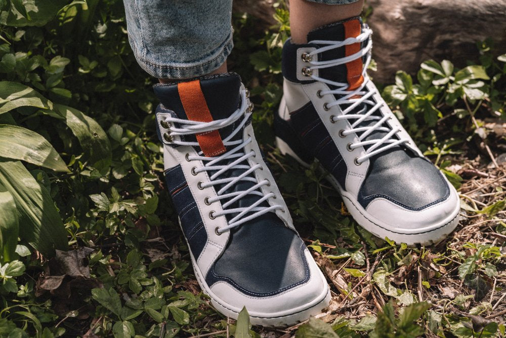 Nahaufnahme eines Paars wasserdichter blau-weißer MONTBLANQ High-Top-Sneaker von ZAQQ, gefertigt aus hochwertigem Nappaleder mit weißen Schnürsenkeln und in einer Mischung aus Marineblau, Weiß und Orange. Sie sind als Outdoor-Schuhe konzipiert und werden von einer Person getragen, die auf Gras inmitten üppigen Grüns steht.