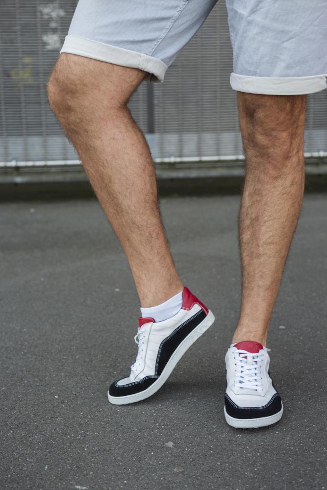 Eine Person trägt die schwarz-weißen QAMPION-Sneakers von ZAQQ, dazu weiße Socken und helle Jeansshorts und steht auf einer Betonfläche mit einem Maschendrahtzaun im Hintergrund. Die Sneaker haben schwarze und rote Akzente und ein flexibles Sohlendesign.