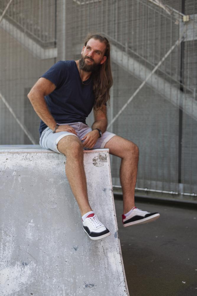 Ein Mann mit langen Haaren und Bart sitzt auf einer Betonrampe. Er trägt ein marineblaues T-Shirt, hellgraue Shorts und die schwarz-weißen ZAQQ QAMPION-Sneaker. Er steht im Freien, im Hintergrund ist ein Metallzaun zu sehen.