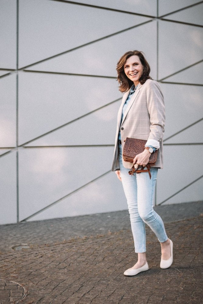 Eine Person steht lächelnd im Freien, trägt einen hellen Blazer und Jeans, trägt helle QARMA Beige Schuhe von ZAQQ und hält eine braune Clutch. Im Hintergrund ist eine moderne Wand mit geometrischen Mustern zu sehen, während der Boden mit Kopfsteinpflaster gepflastert ist.