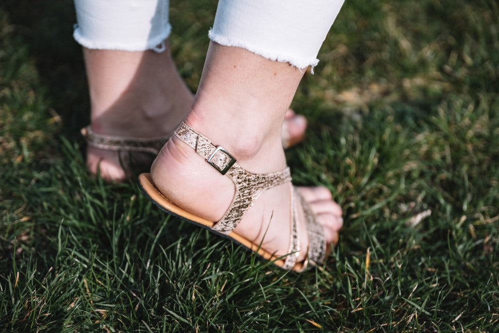 Eine Person in weißer Hose mit ausgefranstem Saum und QEE-Taupe-Sandalen von ZAQQ in modernem Design steht auf grünem Gras. Die glitzernden Sandalen im Barfuß-Sandalen-Stil haben eine seitliche Schnalle und eine flexible Sohle.