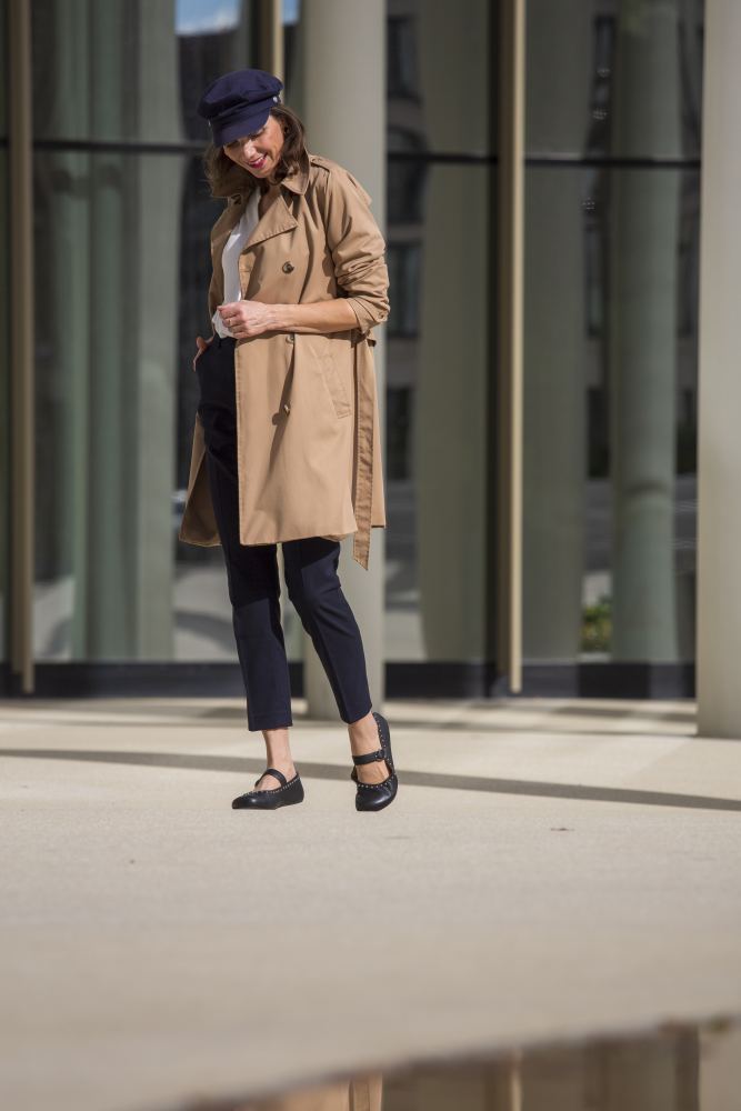 Eine Person in einem beigen Trenchcoat, einer marineblauen Mütze und schwarzen Hosen steht draußen in eleganten ZAQQ DIEQO Nappa Black-Schuhen. Sie lächelt, während sie nach unten blickt, mit einem modernen Gebäude im Hintergrund, das natürliche Fußbewegung auch im urbanen Stil verkörpert.