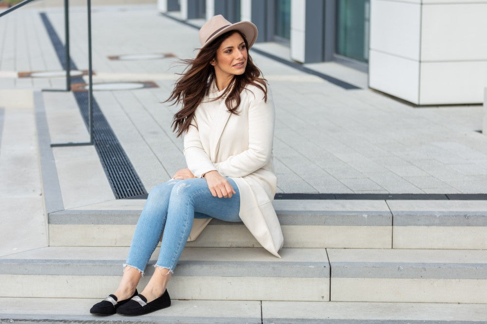 Eine Frau in beigem Mantel, Hut und Jeans sitzt auf Betonstufen. Sie blickt mit einem leichten Lächeln zur Seite. Ihre eleganten Schuhe QERP Velours Black von ZAQQ aus weichem Veloursleder heben sich vor der Kulisse moderner Gebäudefassaden ab.