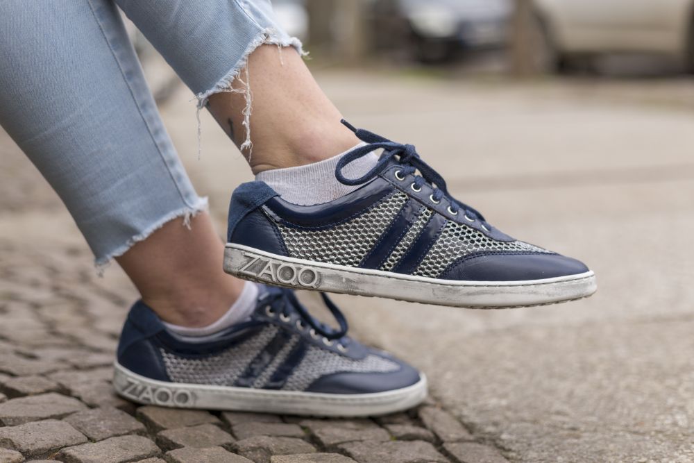 Nahaufnahme einer Person, die QIMBERLY Blue-Sneaker von ZAQQ trägt, kombiniert mit weißen Socken und hellblauen Jeans mit ausgefranstem Saum. Die Schuhe haben eine weiße Sohle mit dem ZAQQ-Logo an der Seite, das die Essenz von Premium-Nappaleder einfängt. Die Person steht draußen auf einem Gehweg aus Kopfsteinpflaster und Beton.