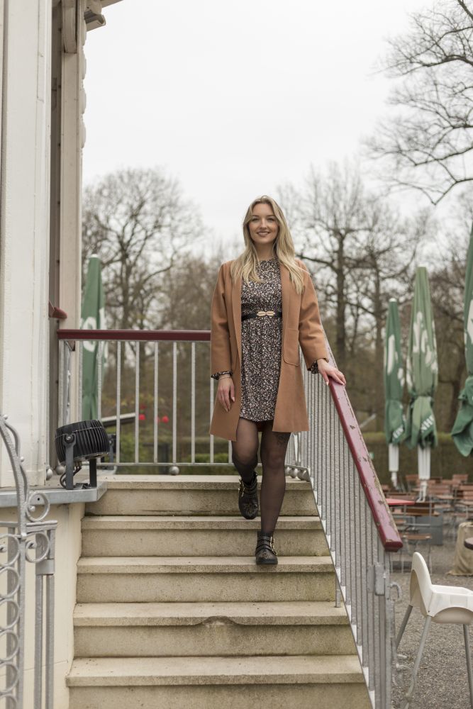 Eine Frau mit langen blonden Haaren, einem braunen Mantel und einem gemusterten Kleid geht selbstbewusst in stilvollen QLANCE Black Gold-Stiefeletten von ZAQQ eine Außentreppe hinunter. Es ist ein bewölkter Tag und im Hintergrund sind Bäume und geschlossene grüne Sonnenschirme in der Nähe eines Gebäudes zu sehen.